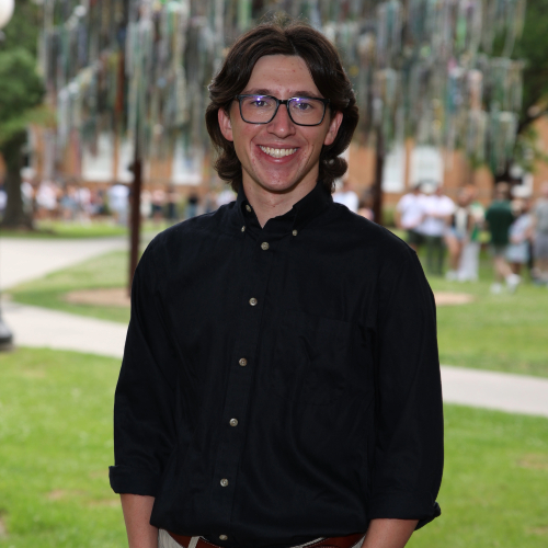 Smiling young man with dark hair and glasses in a black shirt, standing outdoors.
