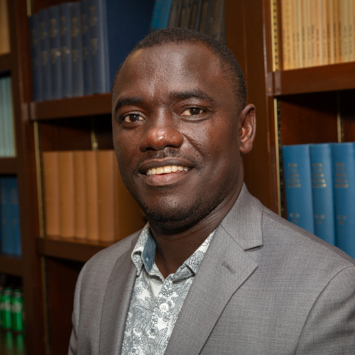 Smiling man in a grey suit and patterned shirt in front of bookshelves.