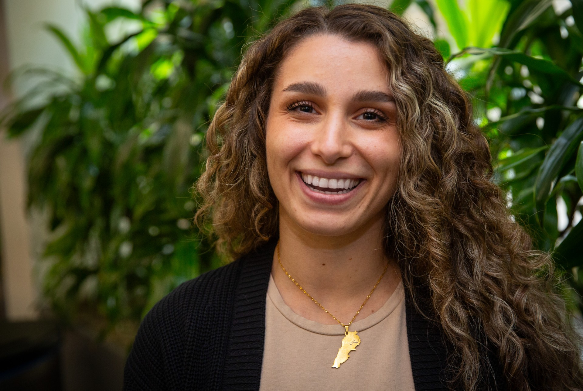 Woman with curly hair smiling, wearing a gold necklace, against green plants.