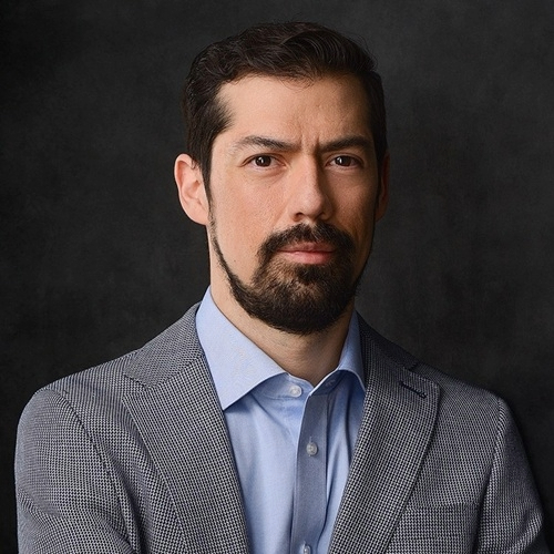 Portrait of a man with dark hair and beard wearing a suit and blue shirt.