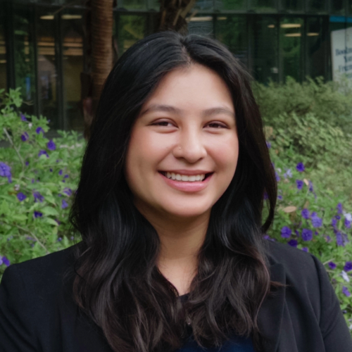 Smiling young woman with long dark hair in a black blazer, outdoors with purple flowers.